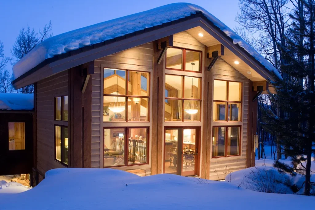 Snow-covered modern wood cabin at twilight with floor-to-ceiling glowing windows