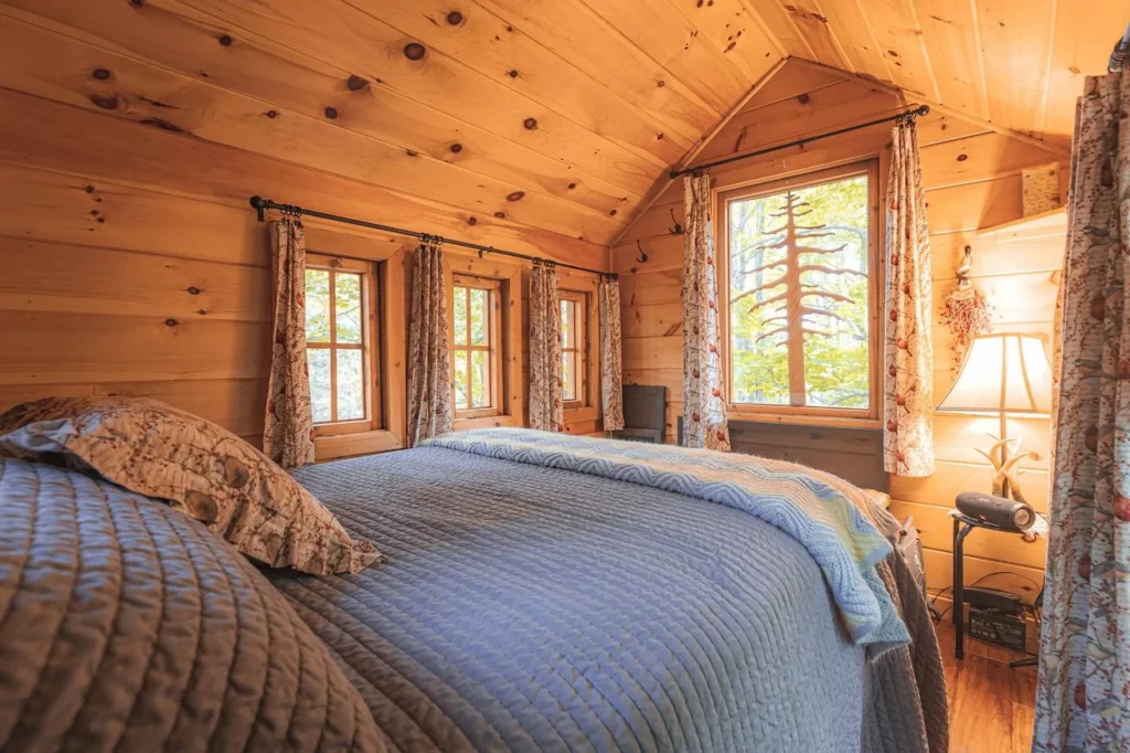 Wood paneled treehouse bedroom featuring a large bed with a grey quilted blanket and pattern curtains