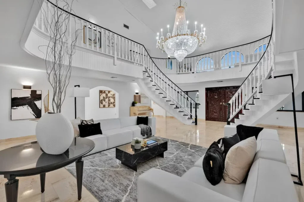 A grand white foyer featuring a large crystal chandelier, a split staircase, and two modern white sofas