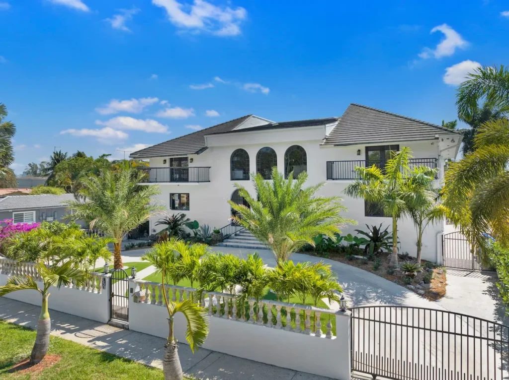 A large three-story white mansion with arched windows, balconies, palm trees, and a gated concrete driveway