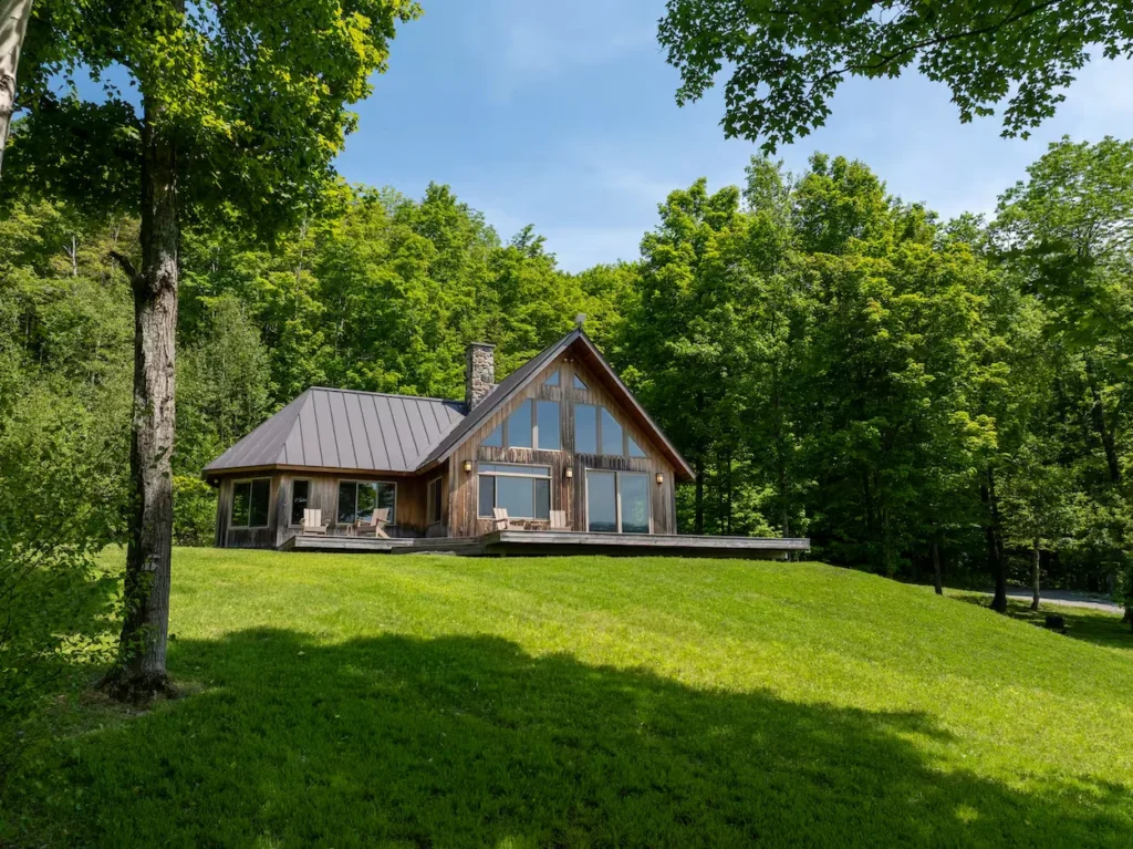 Wooden chalet cabin with a stone chimney and wrap-around deck on a green grassy hill