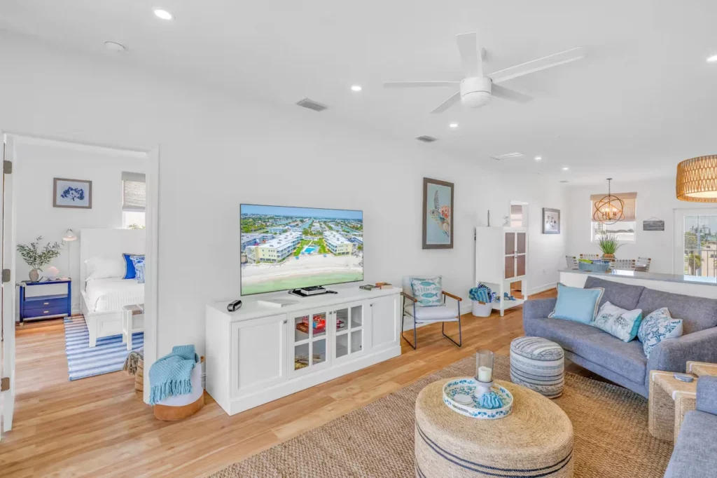 Bright coastal living room with a large TV, grey sofa, round woven coffee table, and an open doorway to a bedroom
