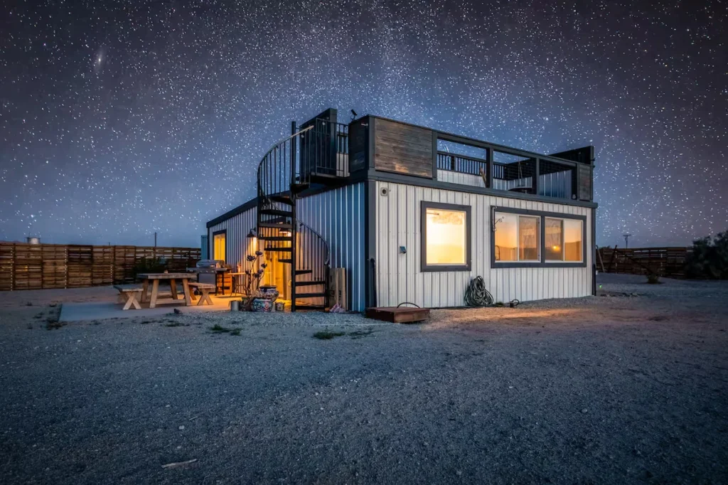 Modern container home at night under a starry sky with an outdoor spiral staircase leading to a rooftop deck