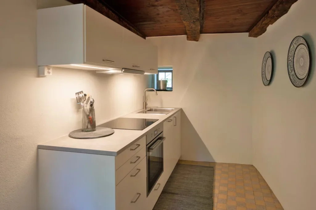 Minimalist white kitchenette with an integrated oven and cooktop under rustic wooden ceiling beams