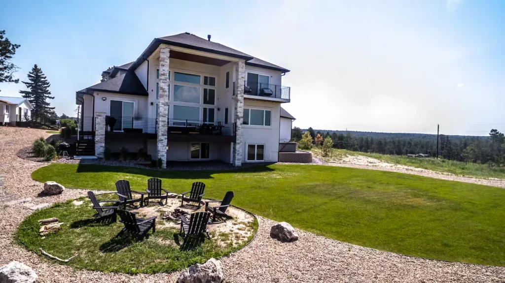 Large two-story modern house with a green lawn and a circular gravel fire pit area with chairs