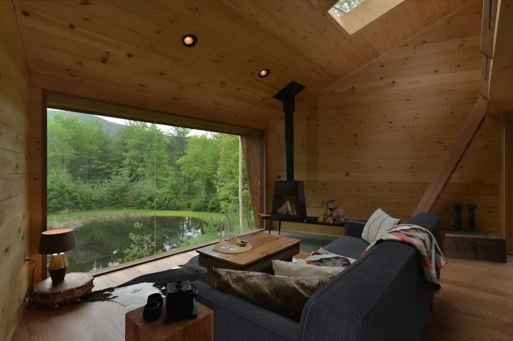 Modern wood cabin living room with a couch, fireplace, and huge window looking at a pond