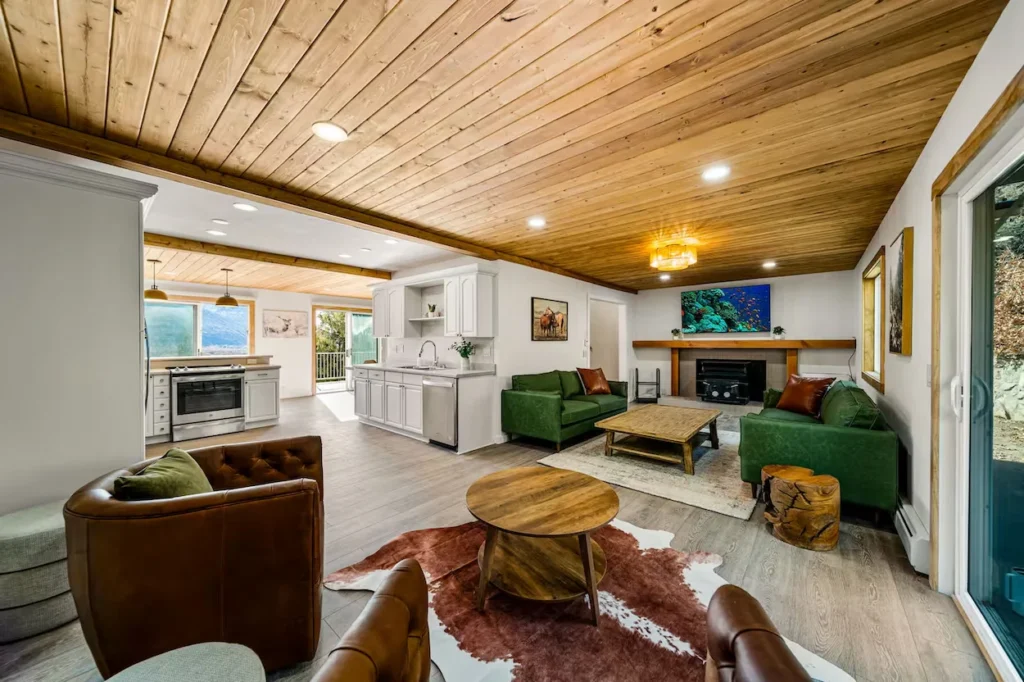 Open-concept cabin living room with green sofas, leather armchairs, a cowhide rug, and white kitchen cabinets