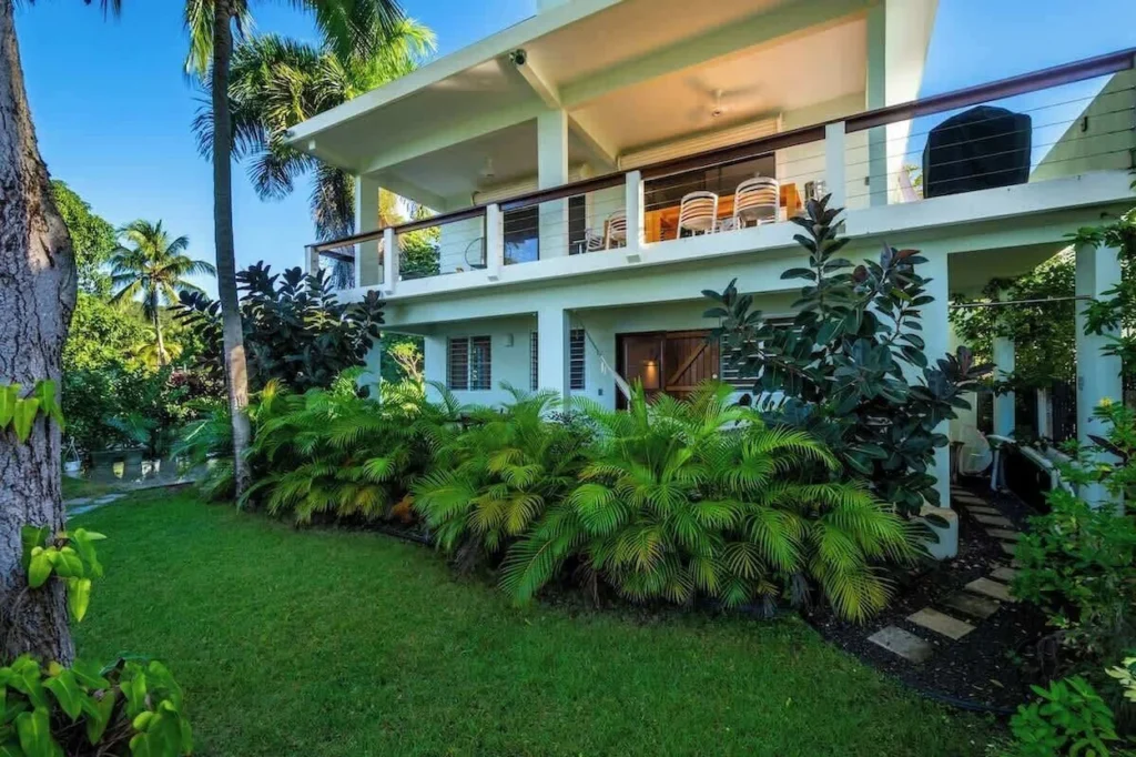 A two-story white modern villa with a wrapping balcony, manicured lawn, and vibrant green palm plants
