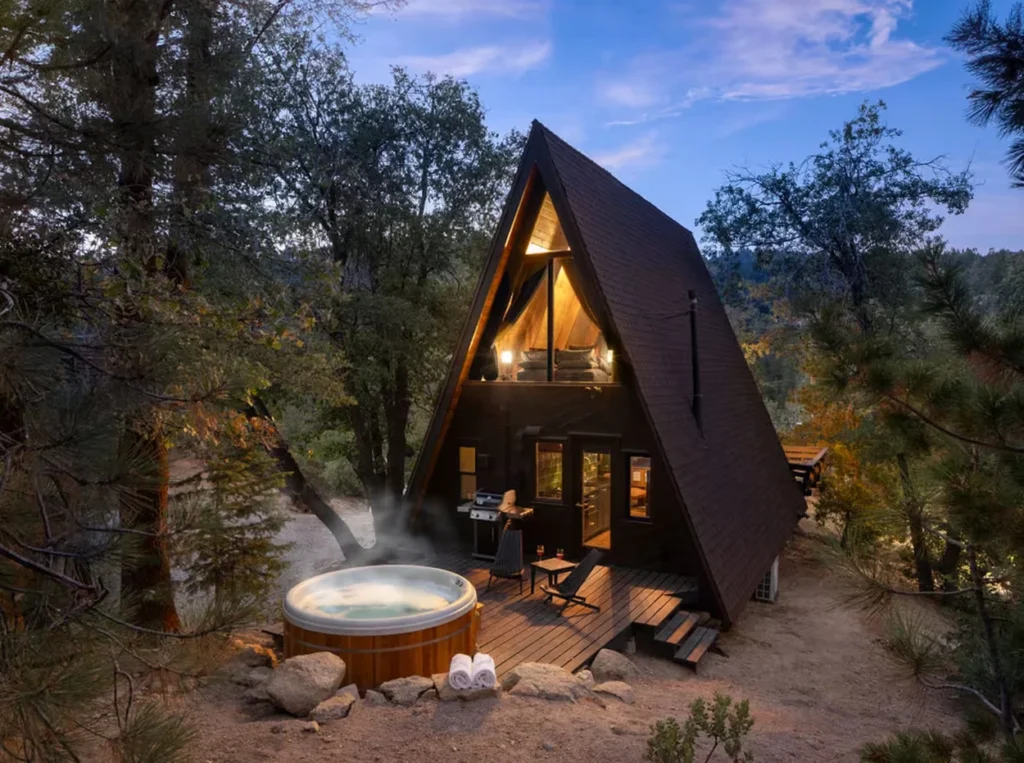 Illuminated A-frame cabin at dusk with a steaming outdoor hot tub on a wooden deck surrounded by trees
