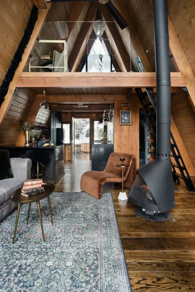 A-frame cabin interior featuring a black freestanding fireplace, a brown chaise lounge, and a loft overhead