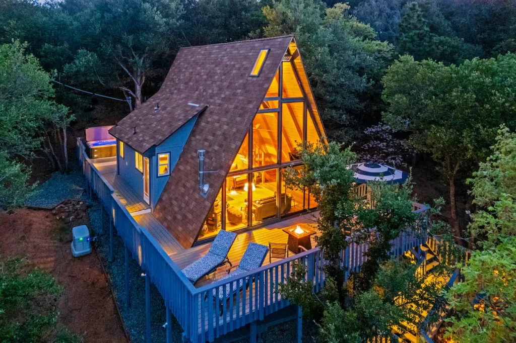 Illuminated A-frame cabin at dusk with a hot tub on the deck