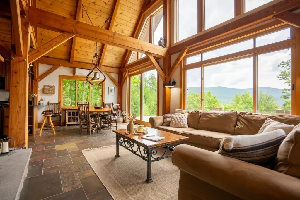 Timber frame cabin interior with a slate tile floor, brown sectional sofa, dining set, and large windows overlooking mountains