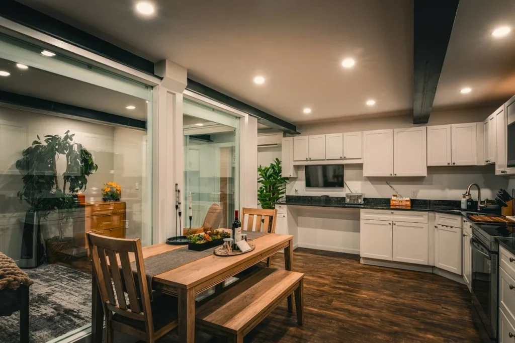 Mirror house kitchen with white cabinets and a wooden dining set next to glass walls