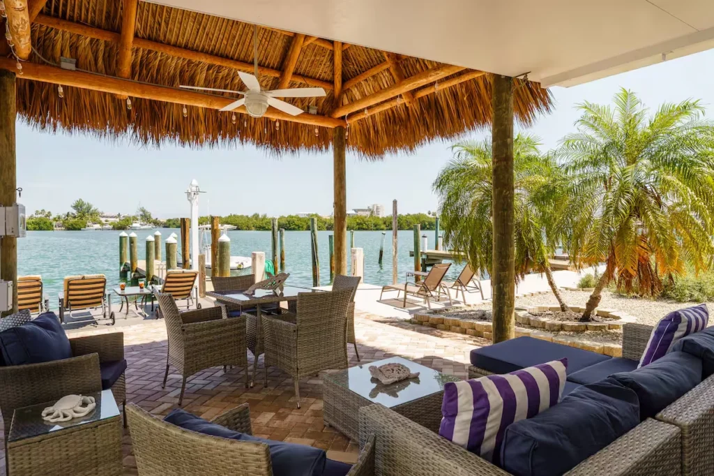 Covered tiki hut patio with wicker lounge furniture and a dining set next to a waterfront dock with palm trees