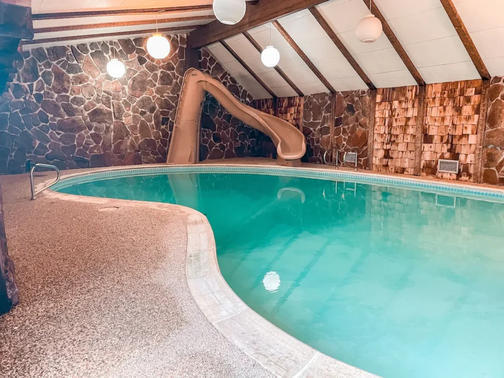Indoor swimming pool with an attached beige water slide, a stone accent wall, and a wood-beamed ceiling