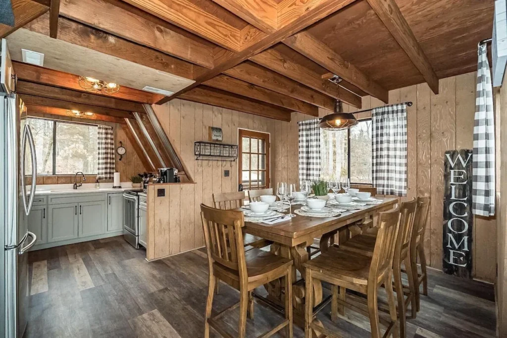 A wooden dining table set for dinner next to a kitchen inside a rustic A-frame cabin