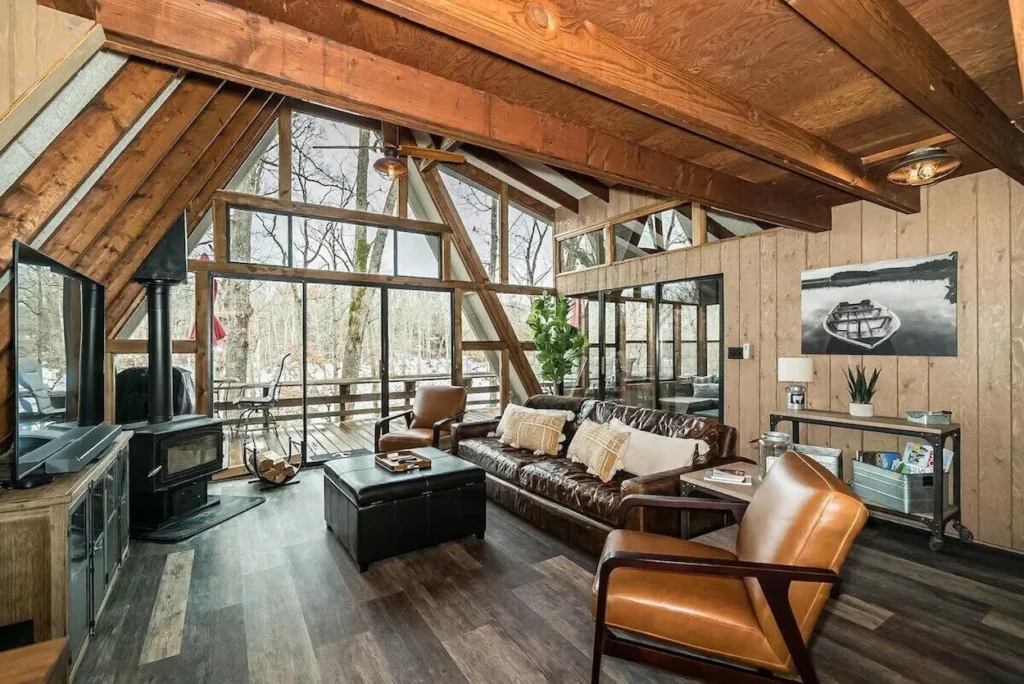 An A-frame cabin living room with leather furniture, a wood-burning stove, and a wall of windows looking outside