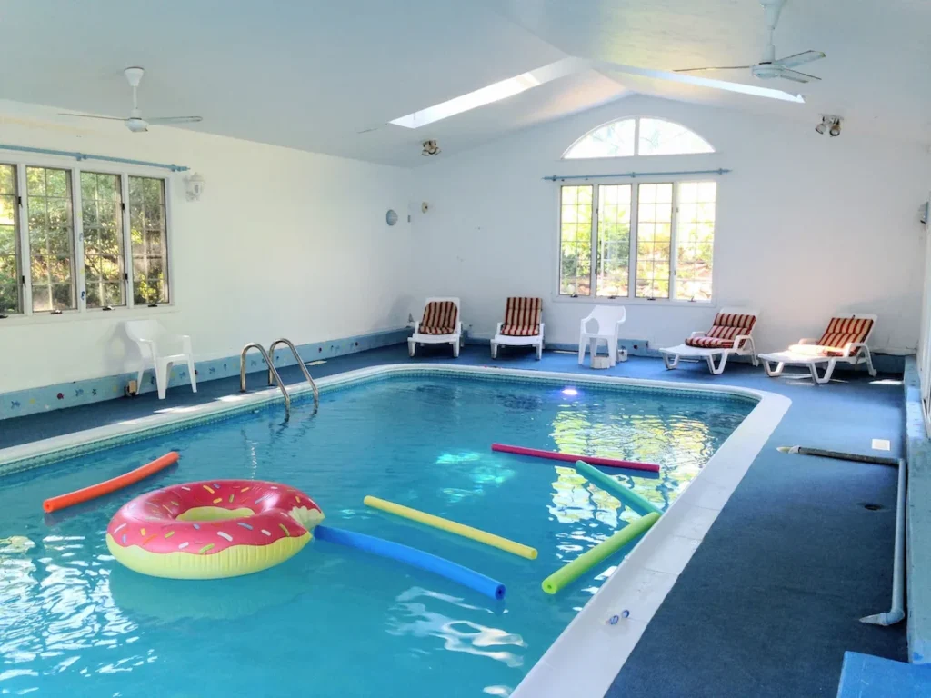 Spacious indoor pool room with a skylight, pool noodles, a donut float, and lounge chairs