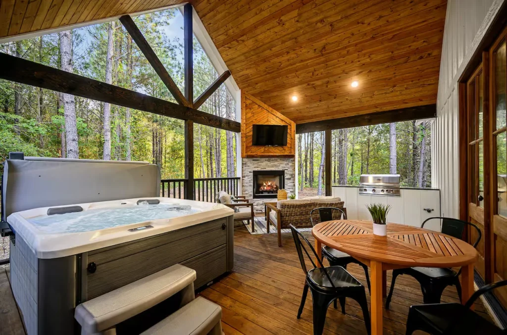 Covered wooden cabin deck with a hot tub, outdoor fireplace, seating area, and dining table facing trees