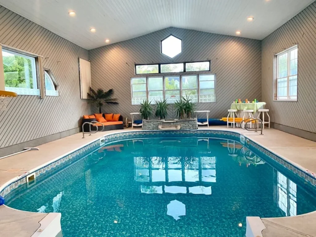 Large indoor swimming pool with blue water, vaulted ceilings, a poolside bar table, and lounge furniture