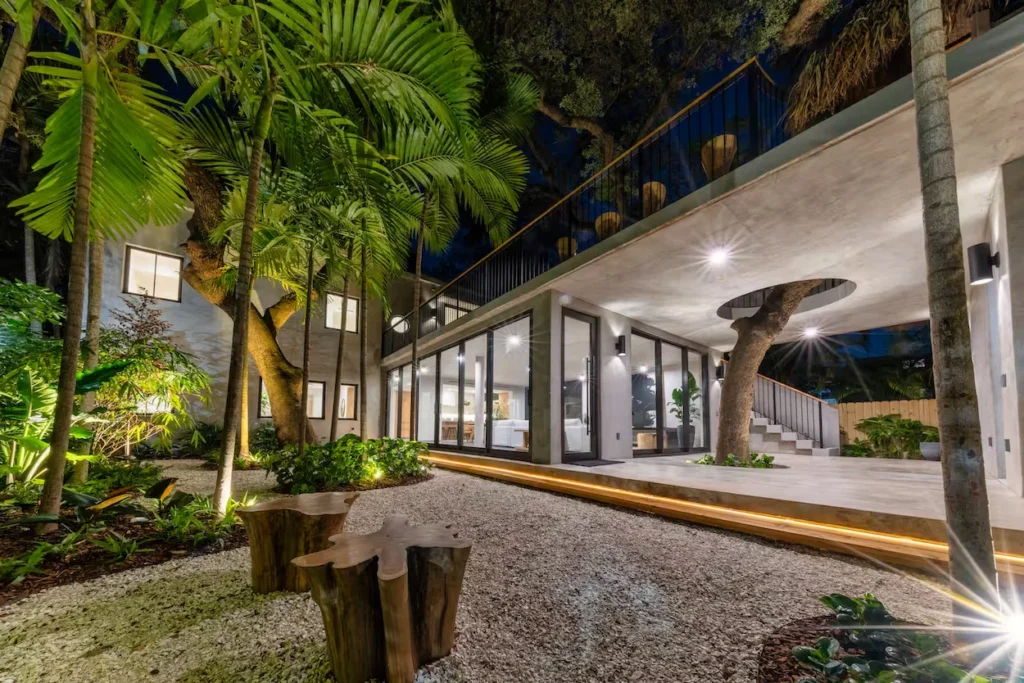 Modern concrete home at night with an illuminated courtyard, palm trees, and a tree growing through the ceiling structure