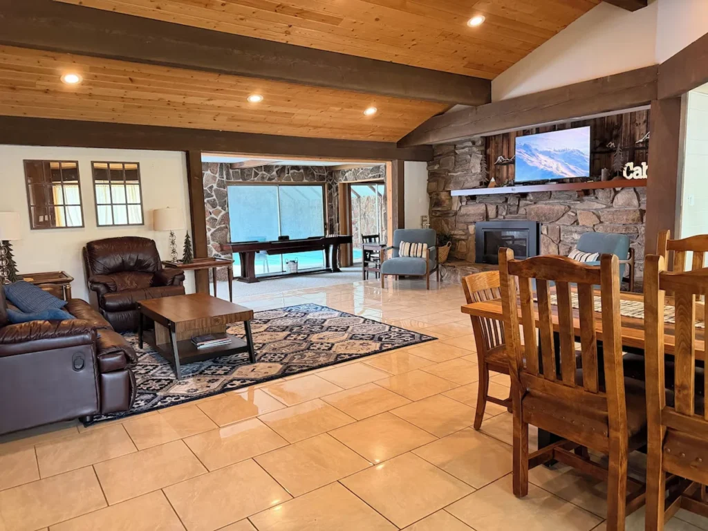 Open-concept living space with a large stone fireplace, wall-mounted TV, leather seating, wood ceilings, and a dining table