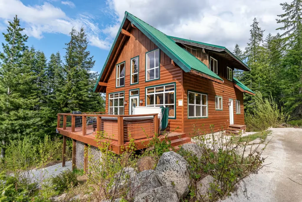 Two-story log cabin with a green roof, large wooden deck, and a hot tub surrounded by pine trees