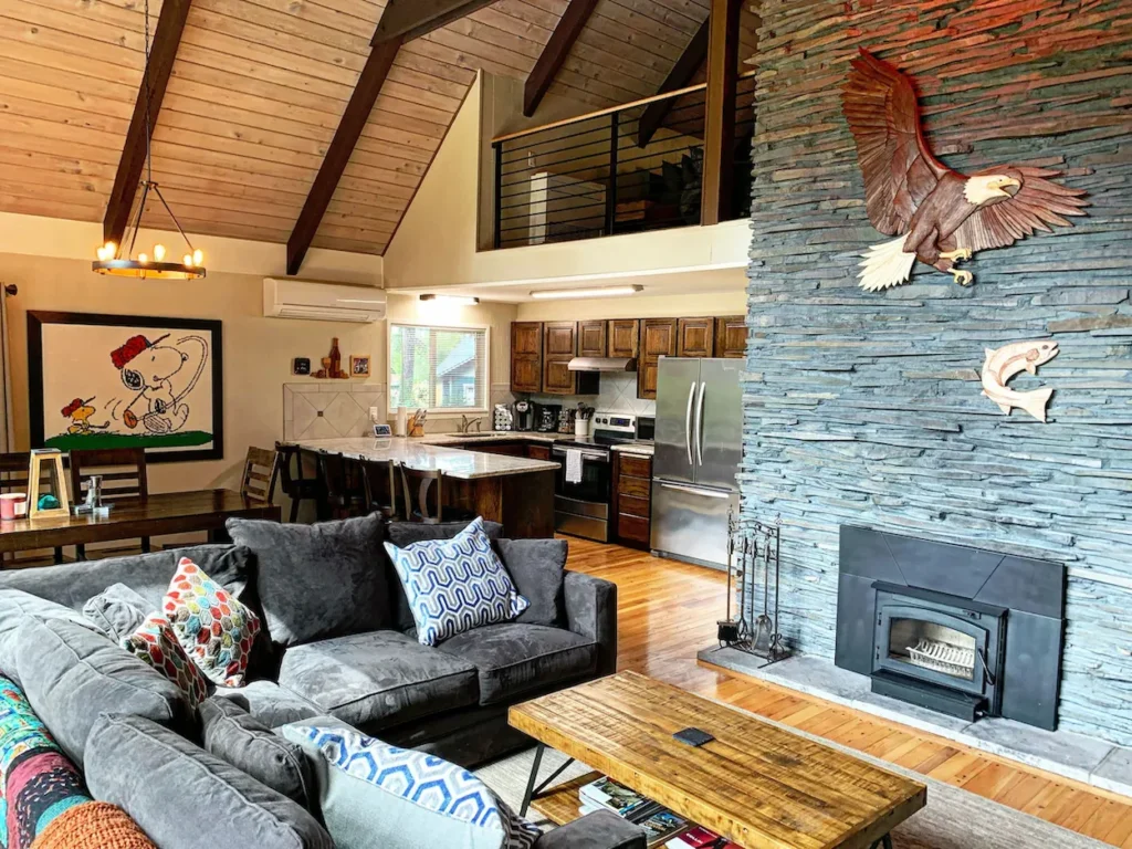 Rustic cabin living room with a large stone fireplace, sectional sofa, open kitchen, and a loft overhead