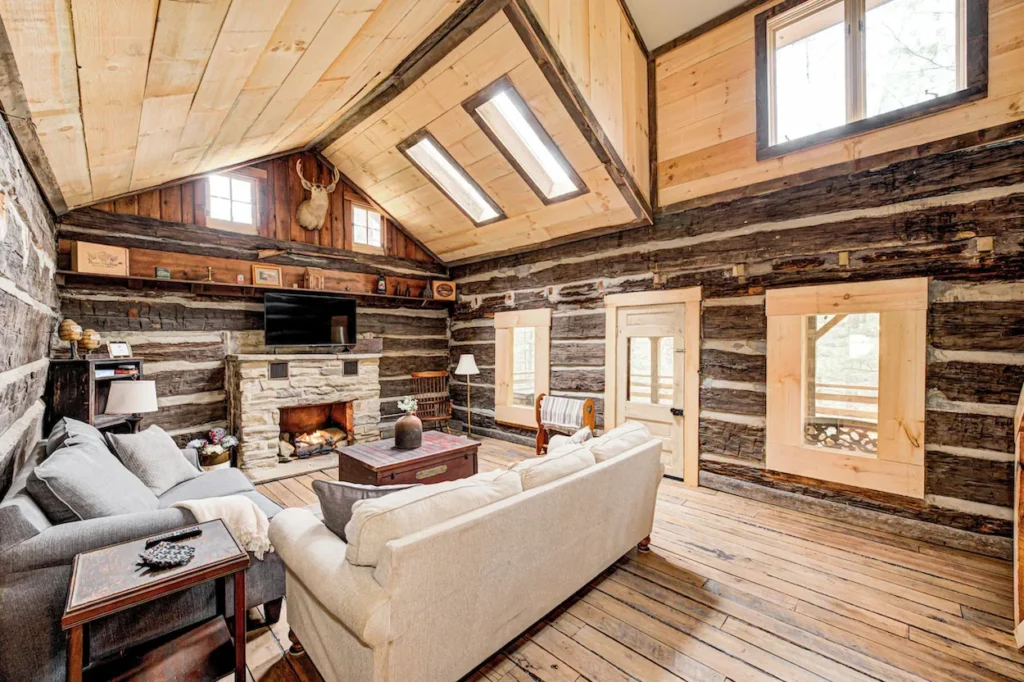 A rustic log cabin living room with light-colored sofas, a stone fireplace, and skylights in a wooden ceiling