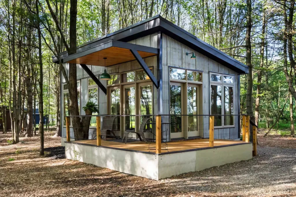 Modern industrial studio cabin with corrugated metal siding, large glass doors, and a front deck surrounded by trees