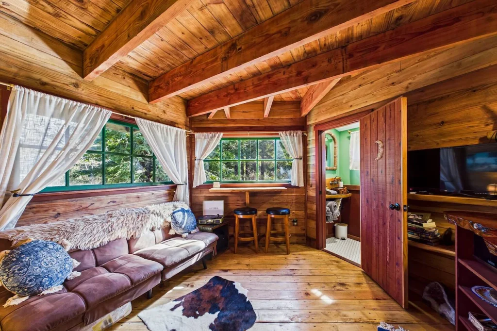 Rustic wood paneled treehouse living room with a brown futon sofa, bar stools, and open door leading to a bathroom