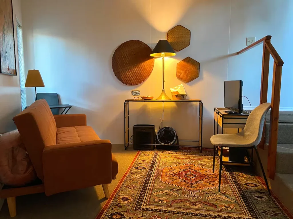 Cozy treehouse corner with an orange futon sofa, a patterned rug, a small desk setup, and a lit table lamp