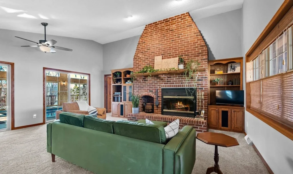 Living room with a large red brick fireplace, green velvet sofa, and wooden built-in shelves