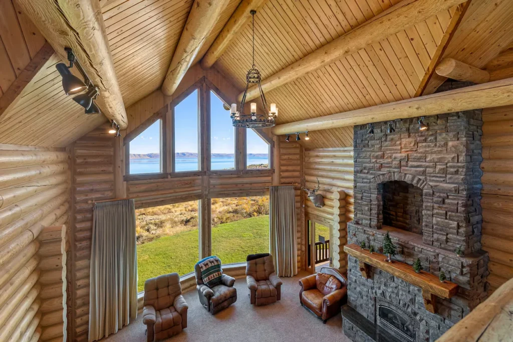 High-angle view of a log cabin living room with a stone fireplace and tall windows facing a lake