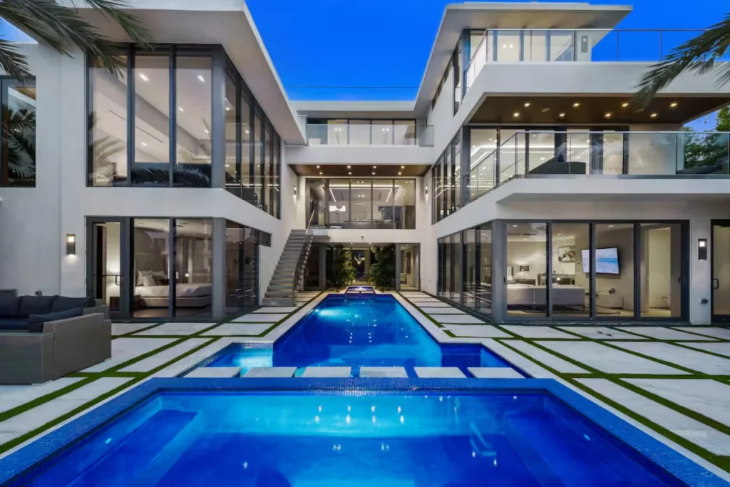 Courtyard of a multi-story modern mansion with a large glowing lap pool at dusk