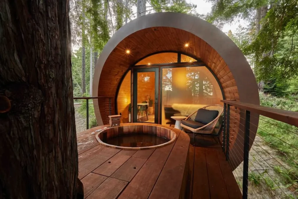 A round barrel style treehouse cabin with a wooden deck, hot tub, and patio chair in the woods