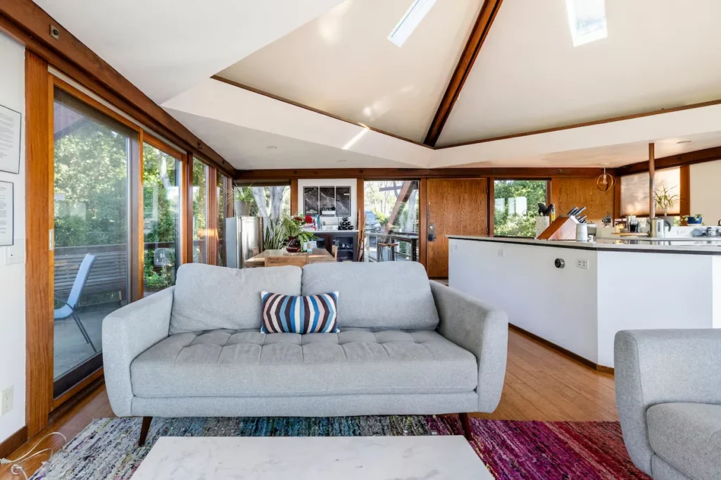 Bright treehouse living room with a gray tufted couch, wooden trim windows, and an open kitchen area