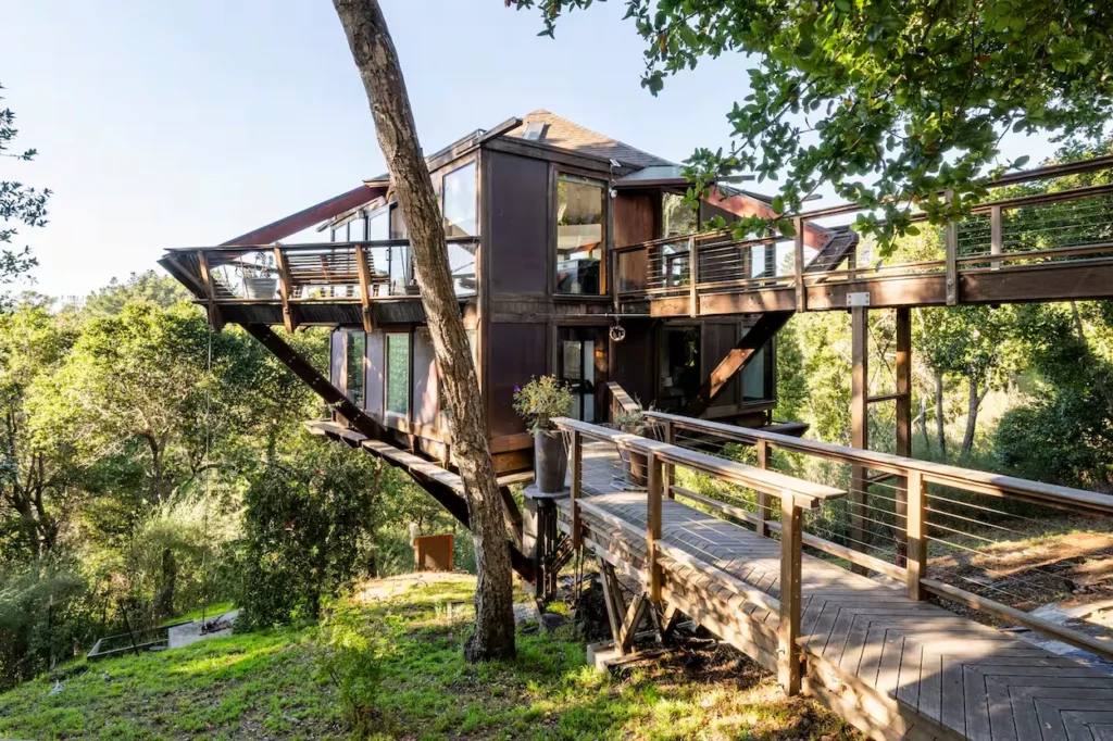 A modern multi-level dark wood treehouse with suspended balconies and wooden walkways on a forested hillside
