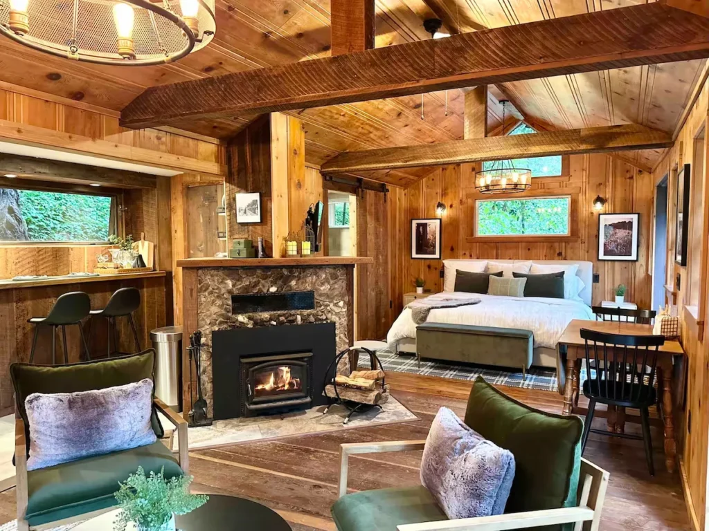 Cozy wood paneled treehouse studio with a stone fireplace, green armchairs, and a bed in the background