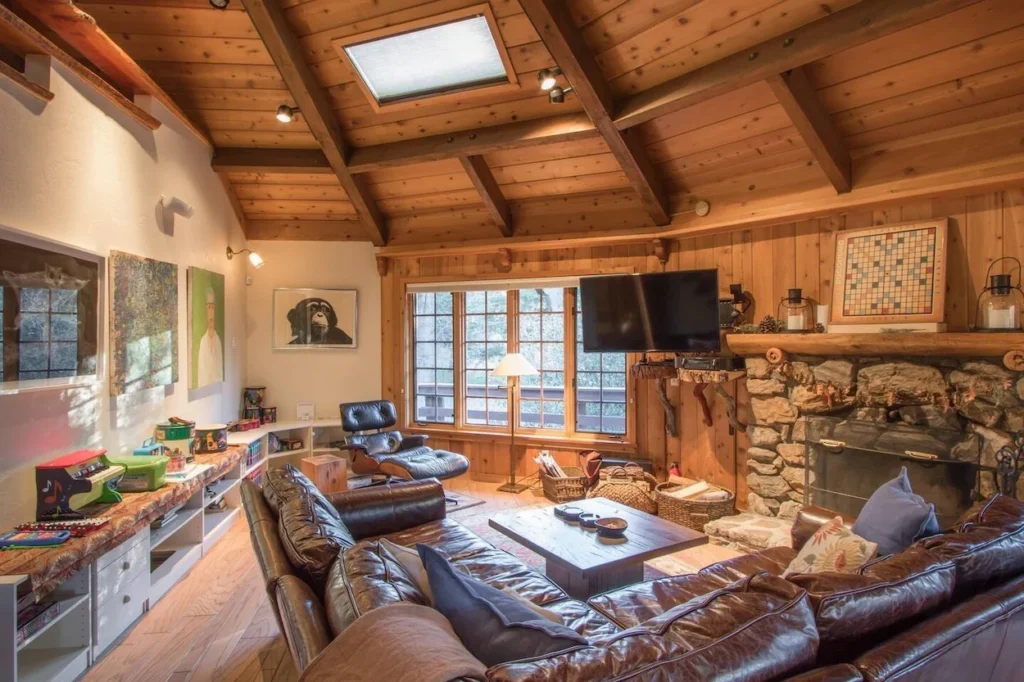Rustic treehouse living room with a stone fireplace, wood-paneled walls, leather sectional, and a skylight ceiling