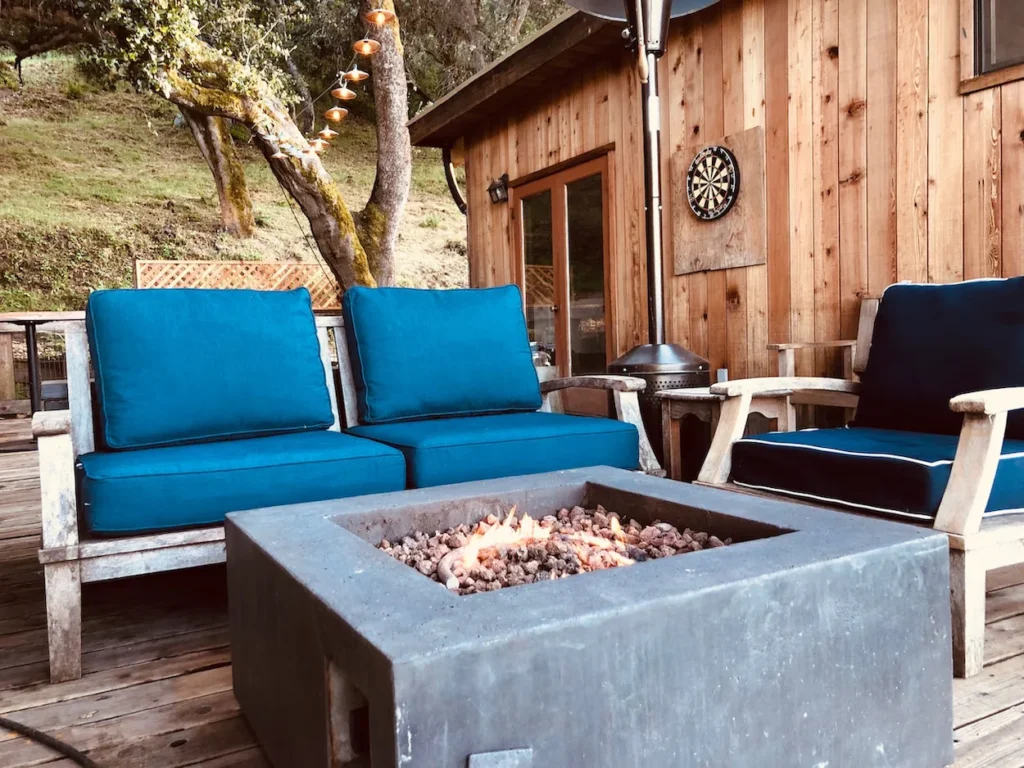 Wooden treehouse deck with blue cushioned chairs arranged around a lit square concrete gas fire pit
