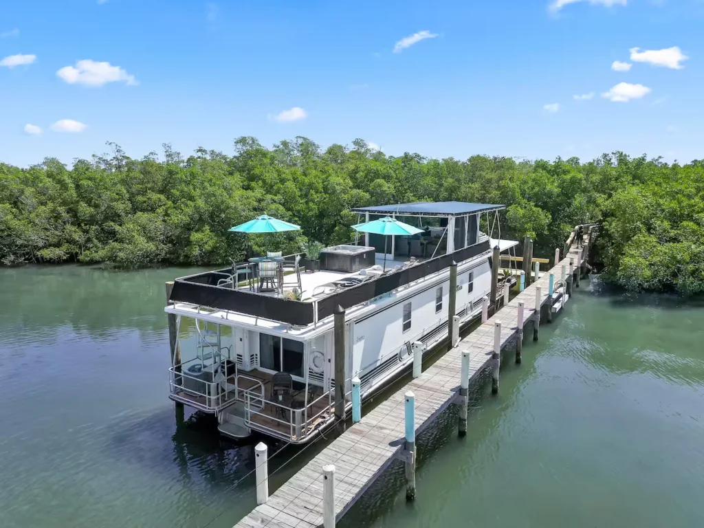 Aerial view of a white multi-story houseboat docked along a wooden pier in a scenic mangrove channel