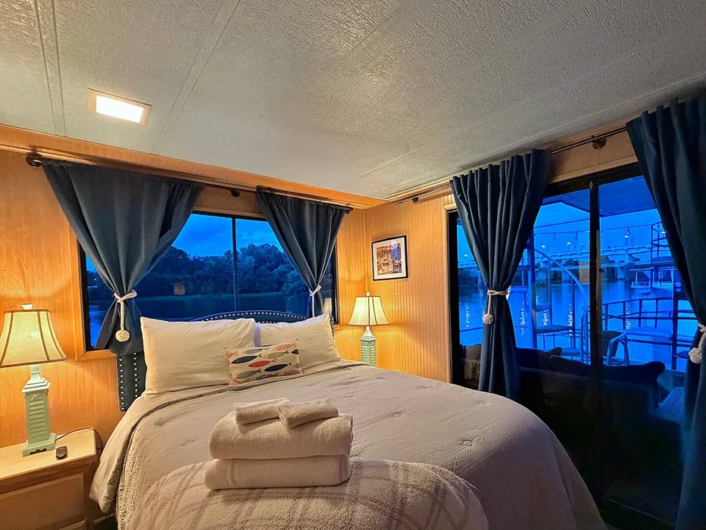 Houseboat bedroom with a bed, nautical lamps, blue curtains, and sliding doors opening to a water deck view
