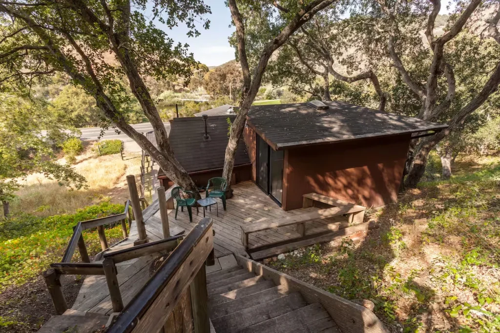 A brown hillside cabin featuring a wooden deck with green chairs and a steep wooden staircase leading down