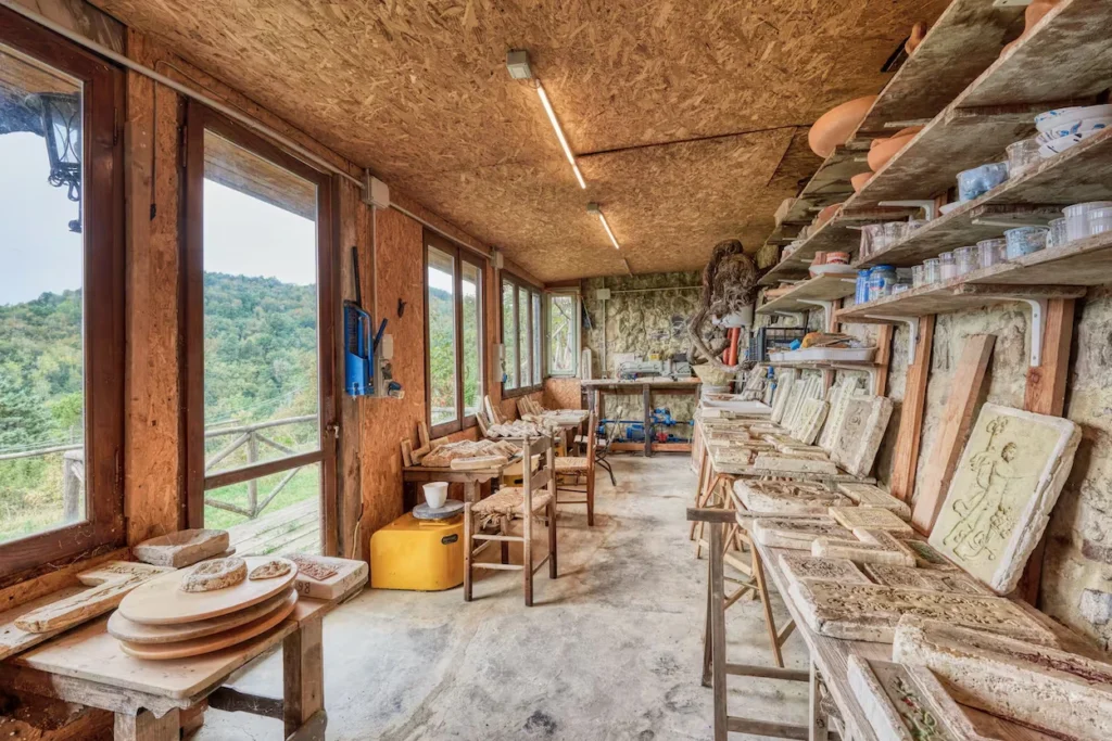 Art studio workshop filled with stone carvings, wooden workbenches, and windows overlooking green hills
