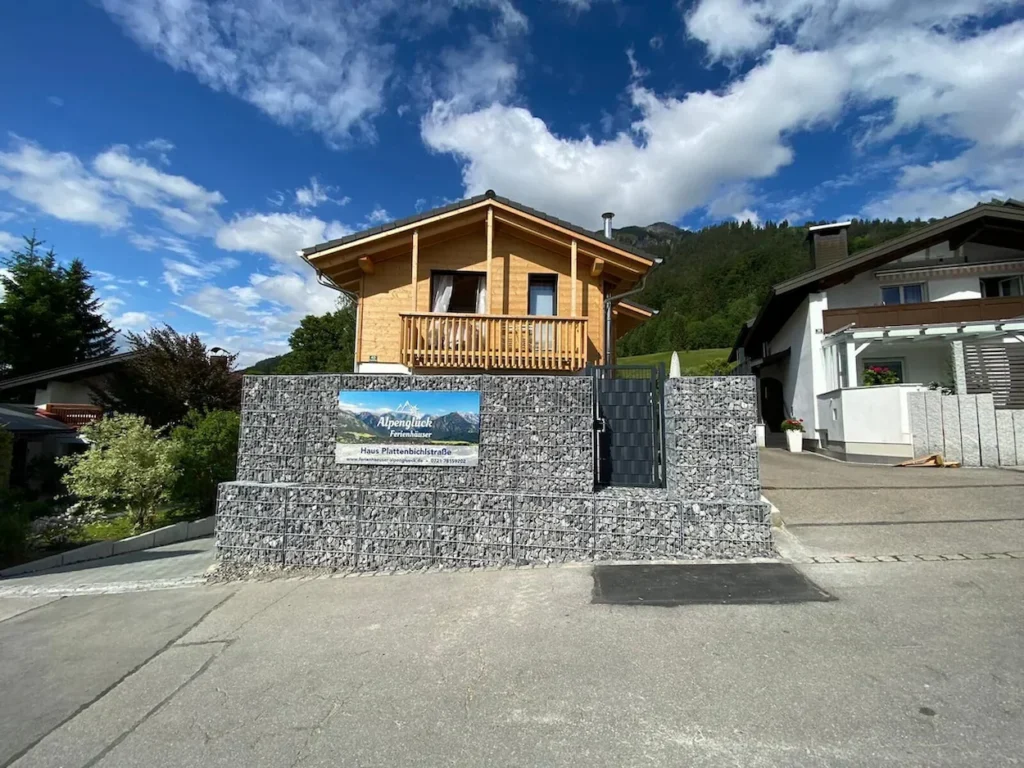 Two-story timber alpine cabin behind a grey stone gabion wall with an Alpenglueck sign