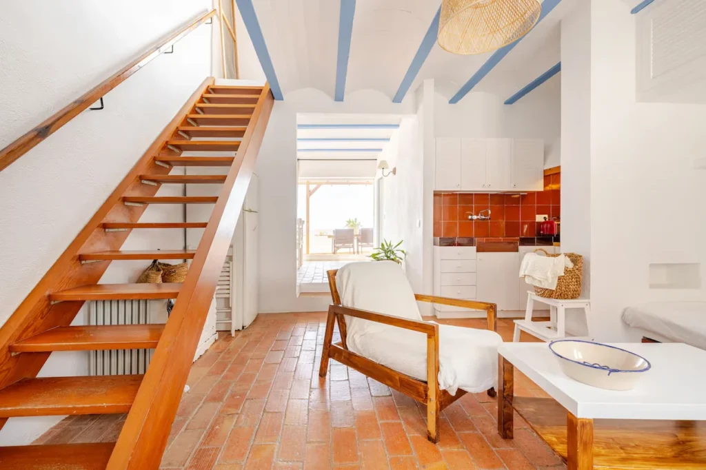 Bright cottage interior with a wooden staircase, terracotta tile flooring, and a white armchair