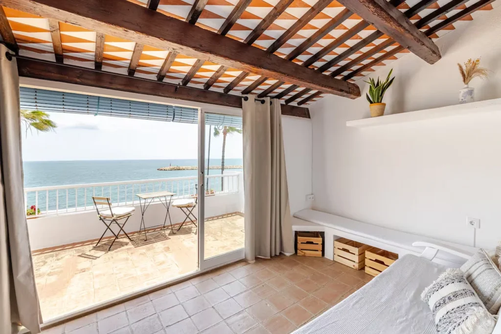 Spanish coastal home interior with wood ceiling beams, opening to a sunlit balcony overlooking the ocean