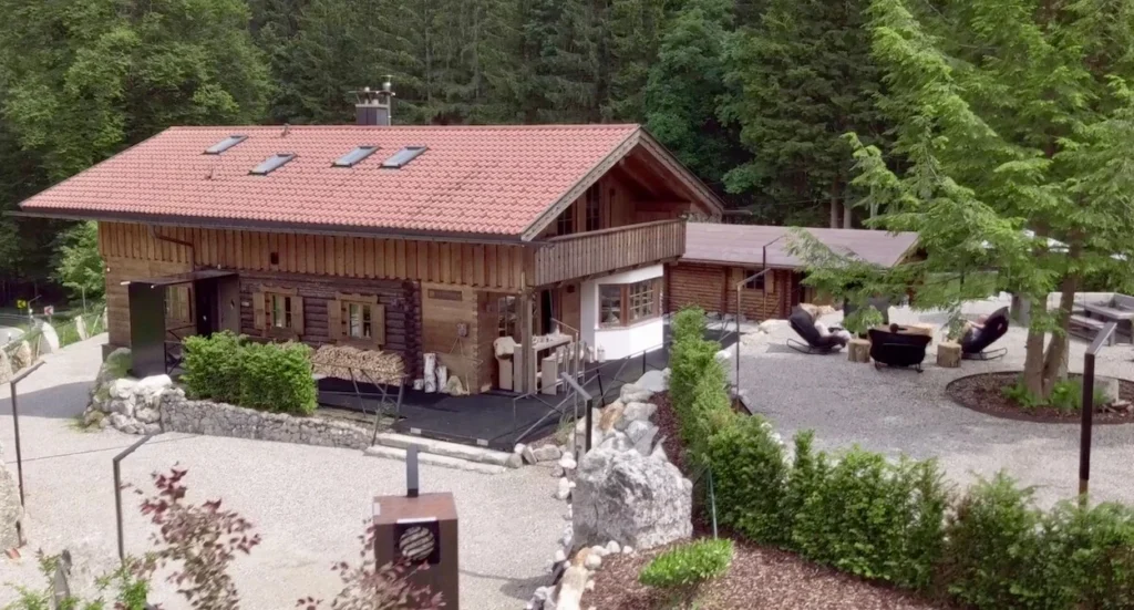 Luxury timber chalet with a red tiled roof, a gravel patio with lounge chairs, and a surrounding pine forest