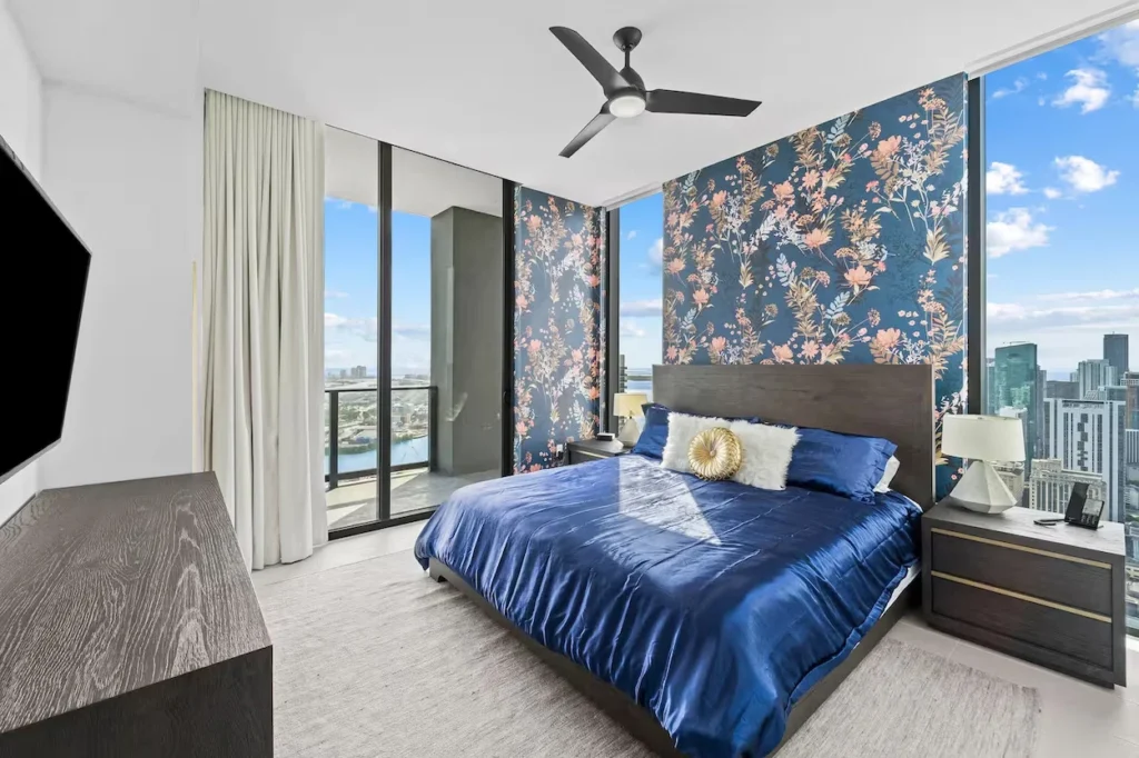 Modern penthouse bedroom with a blue bed, floral accent wallpaper, and large windows overlooking a city